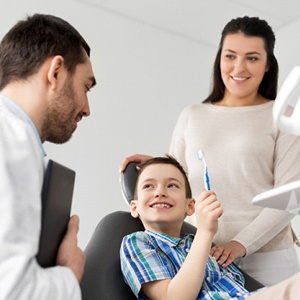  
A dentist talking to a young boy in the dentist’s chair who’s holding a toothbrush as his mother looks on
