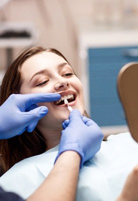 Dentist holding shade guide to woman’s teeth as she looks at reflection in mirror